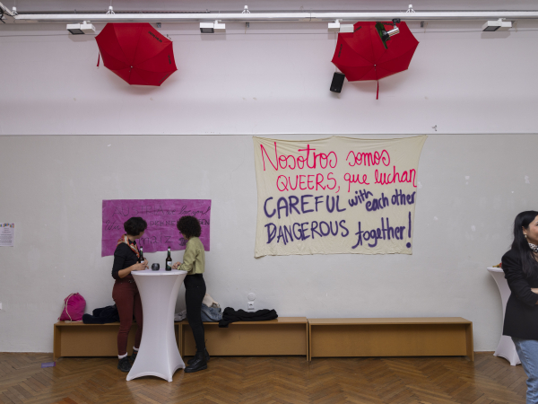 Zwei Menschen im Raum. Hinter ein Banner "Nosotros somos queer que luchan.."